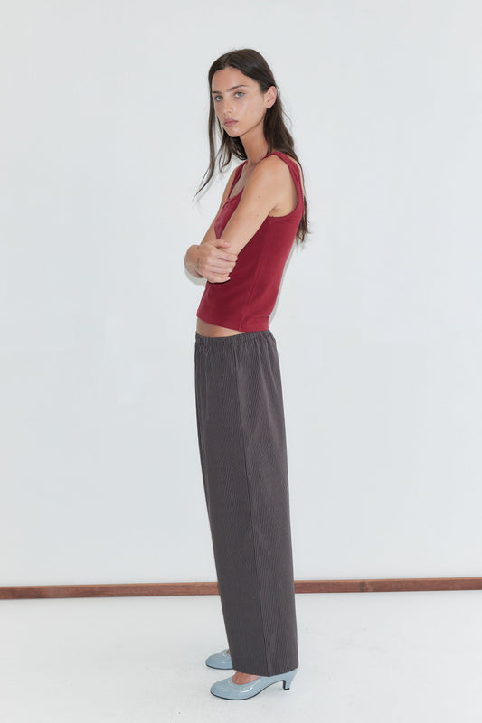 A woman with long dark hair stands in profile against a plain white background wearing the Ease Trouser - Brown Blue Stripe by Deiji Studios, dark brown wide-leg trousers with a fine blue pinstripe, with a red square-neck tank and pale blue heels.
