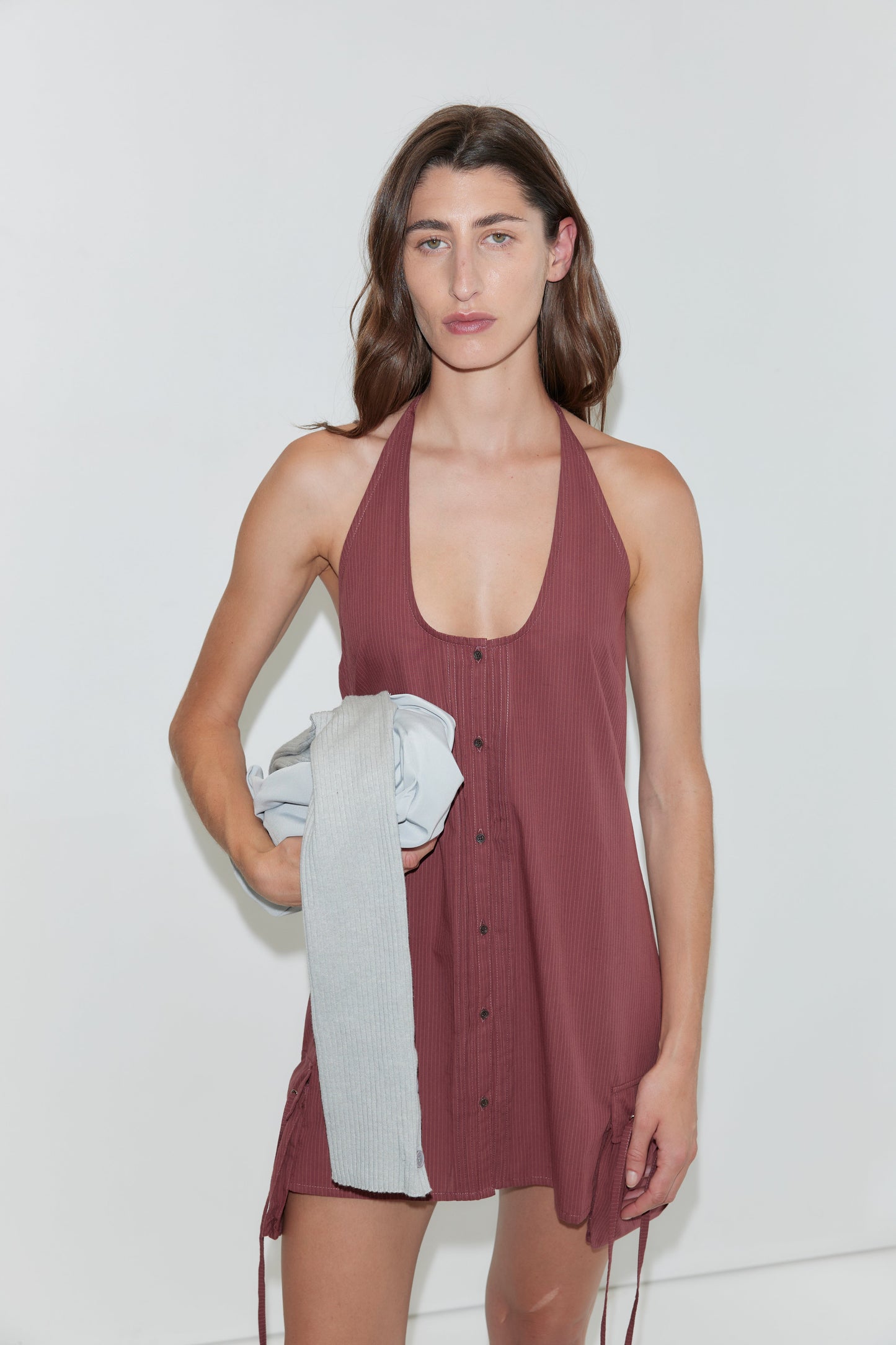 A person with long brown hair, wearing a sleeveless, button-up Pocket Halter Dress in Rust Stripe, stands against a white background holding a folded gray garment in one hand.