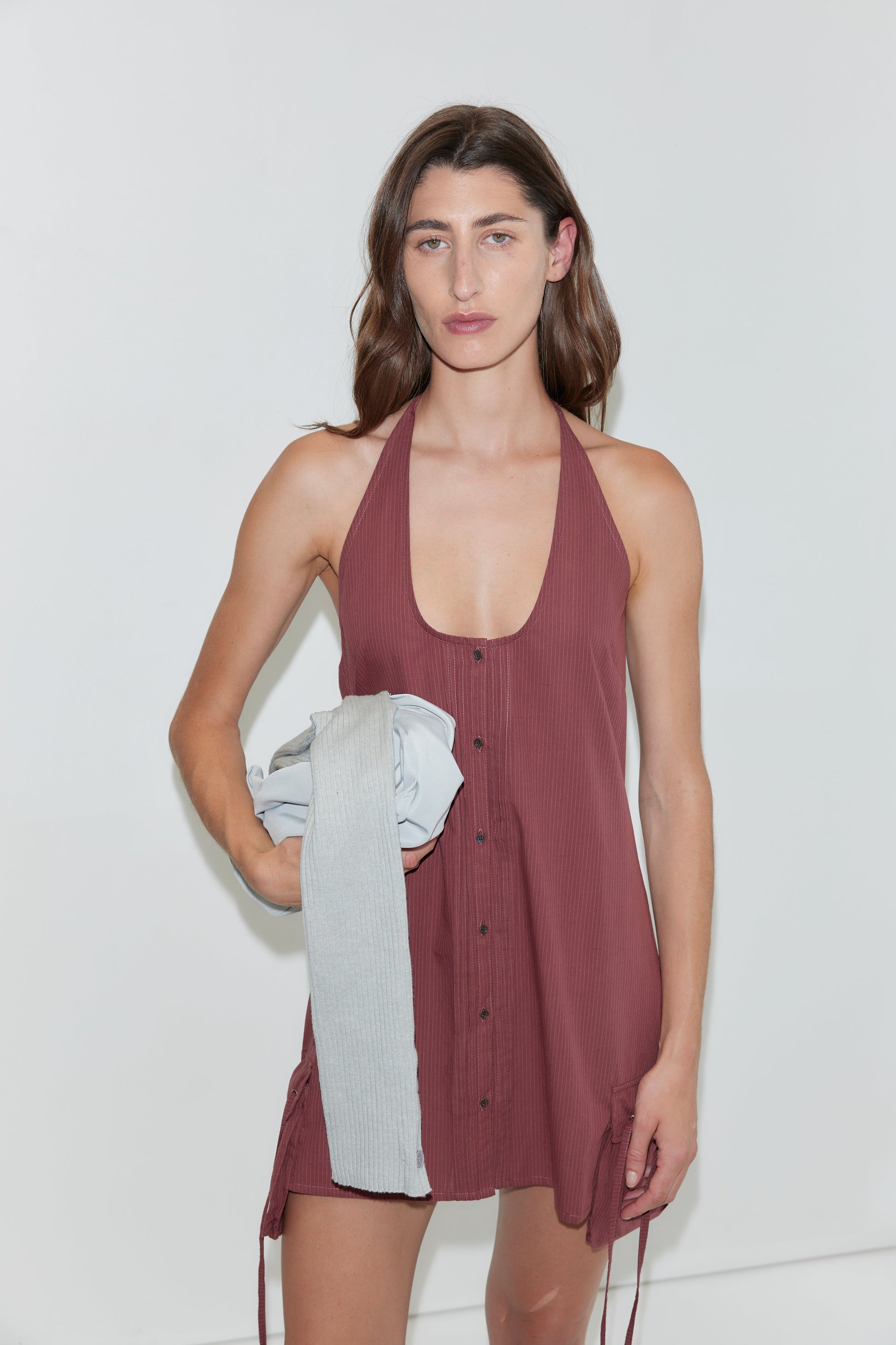 A person with long brown hair, wearing a sleeveless, button-up Pocket Halter Dress in Rust Stripe, stands against a white background holding a folded gray garment in one hand.