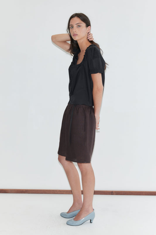 A woman with long dark hair stands in profile against a plain white background wearing the Flounced Top - Black by Deiji Studios, a black top with a frilled square neckline and puff sleeves, with brown striped linen shorts and pale blue heels.