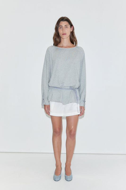 A woman with long dark hair stands against a plain white background wearing the Wided Longsleeve - Grey Marle by Deiji Studios, a grey marle jersey top with a wide neckline and drawstring hem, with white shorts and pale blue heels.