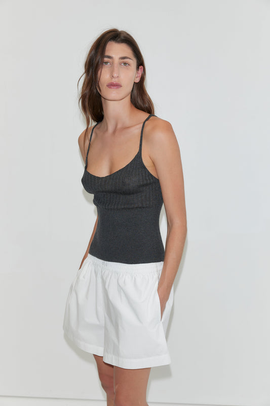 A woman with long brown hair stands against a white background, wearing an Organic Cotton Boxer in white and a fitted black tank top, hands in pockets, looking slightly to the side.