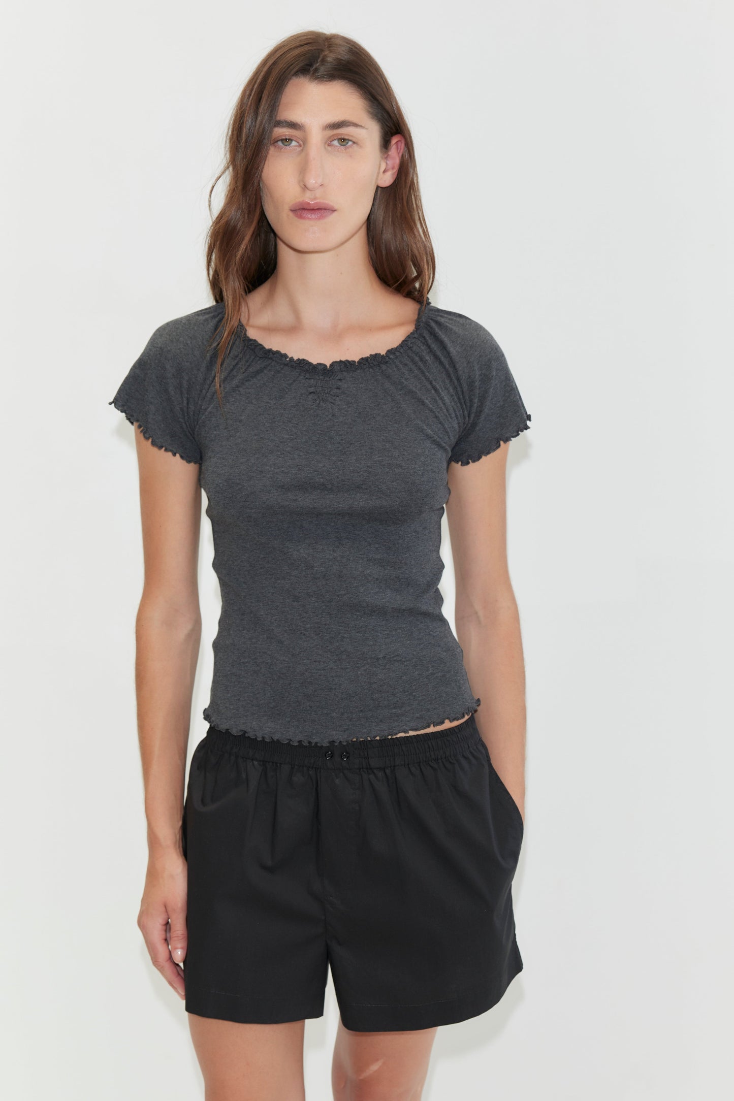 A woman with long brown hair stands against a plain white background, wearing loose black Organic Cotton Boxer shorts and a dark grey fitted short-sleeve top, with one hand in her pocket.