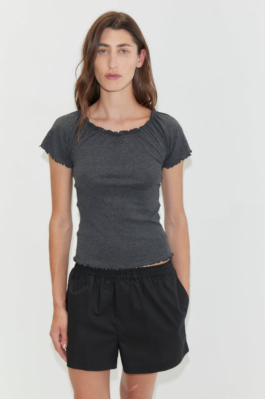 A woman with long brown hair stands against a plain white background, wearing loose black Organic Cotton Boxer shorts and a dark grey fitted short-sleeve top, with one hand in her pocket.