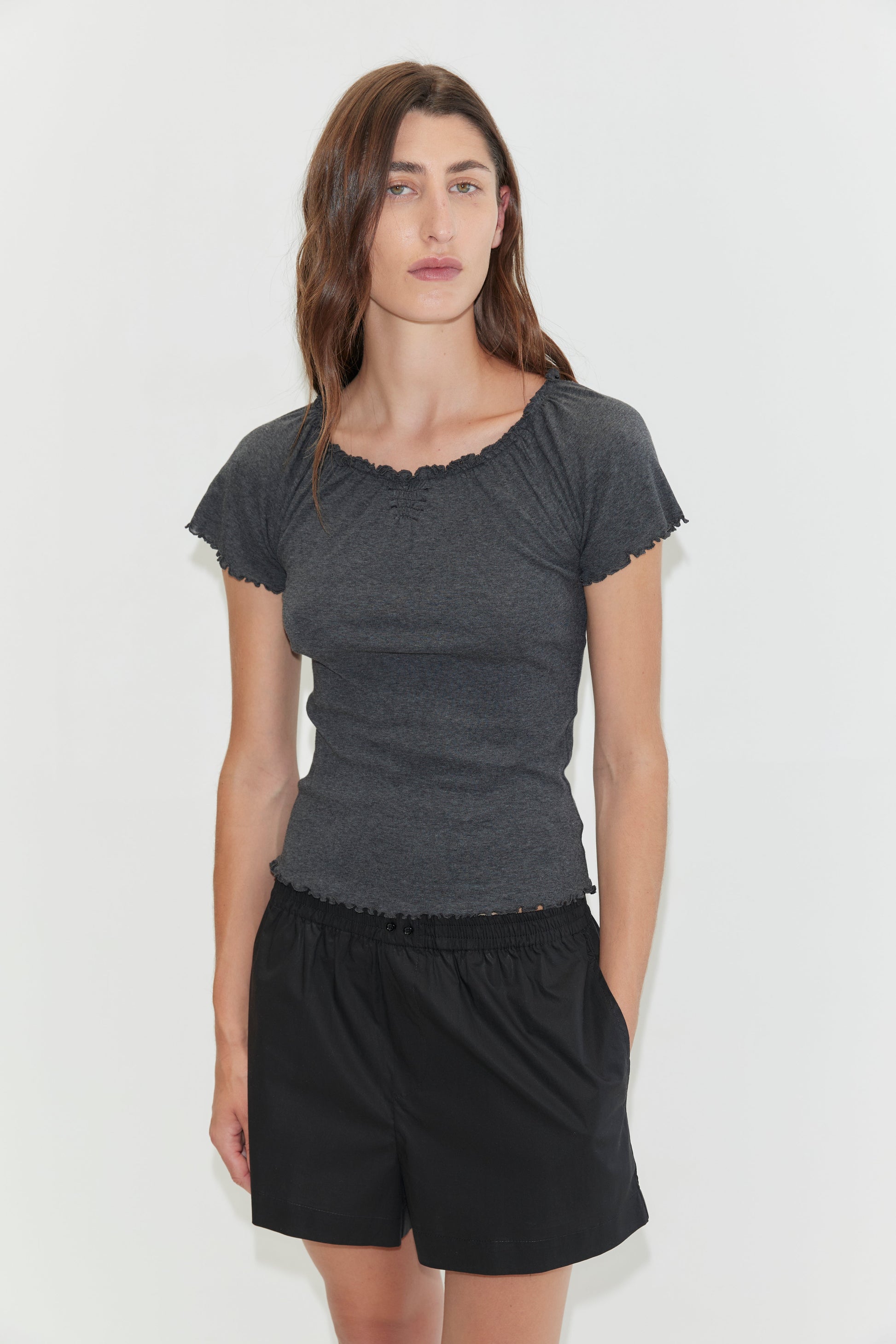 A woman with wavy brown hair wears a fitted dark gray top and loose Organic Cotton Boxer - Black shorts, standing against a plain white background with a neutral expression.