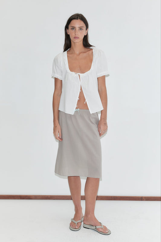 A woman with long dark hair stands against a plain white background wearing an open white frill top and the Basic Skirt - Grey by Deiji Studios, a grey cotton voile midi skirt with a soft elasticated waist, paired with grey flip-flops.