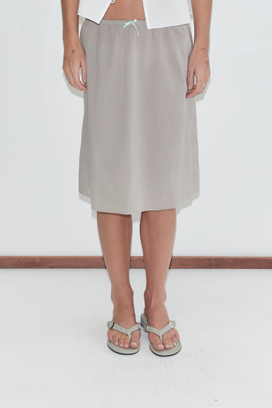 A cropped view of the Basic Skirt - Grey by Deiji Studios, a grey cotton voile midi skirt with a soft elasticated waistband and contrast bow at the centre front, worn with grey flip-flops against a plain white background.