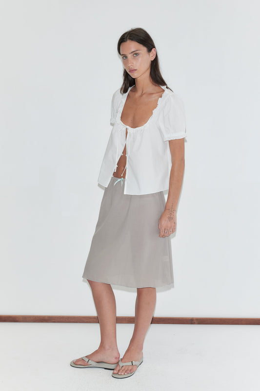 A woman with long dark hair stands against a plain white background wearing an open white frill top and the Basic Skirt - Grey by Deiji Studios, a grey cotton voile midi skirt, with grey flip-flops.
