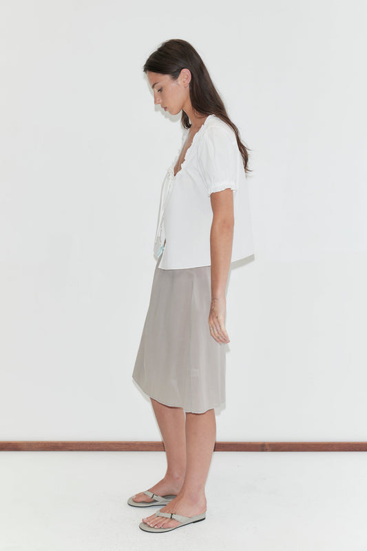 A woman with long dark hair stands in profile against a plain white background, looking down, wearing a white frill top and the Basic Skirt - Grey by Deiji Studios, a grey cotton voile midi skirt, with grey flip-flops.