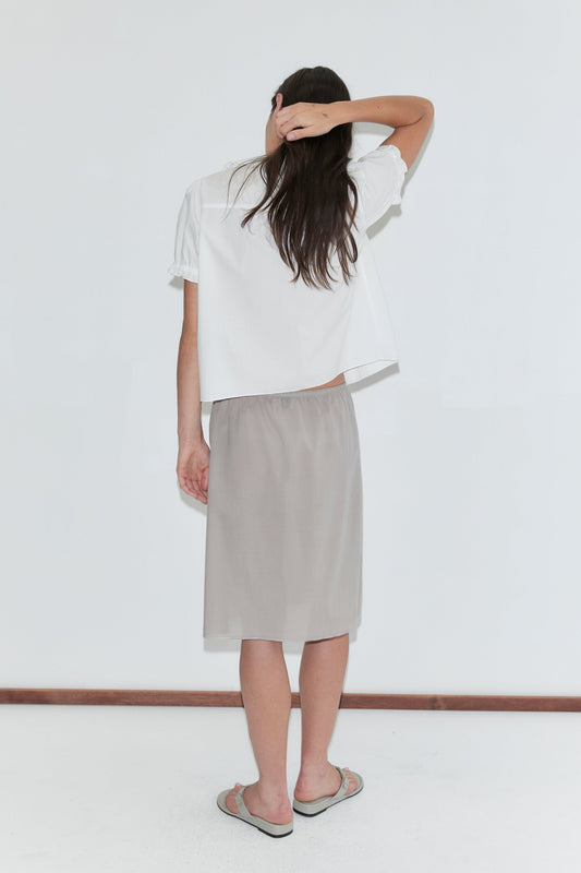 A woman with long dark hair stands with her back to the camera against a plain white background, wearing a white frill top and the Basic Skirt - Grey by Deiji Studios, a grey cotton voile midi skirt, with grey flip-flops.