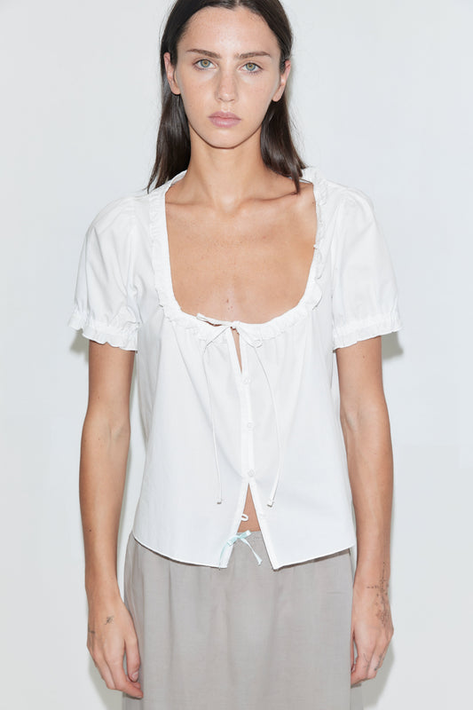 A woman with long dark hair stands facing away against a plain white background with one hand raised to her head, wearing a white frill top and the Basic Skirt - Grey by Deiji Studios with grey flip-flops.