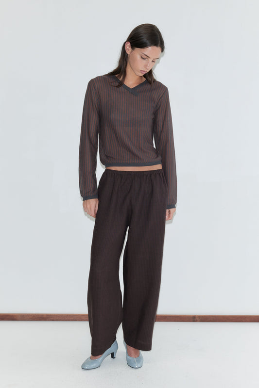 A woman with long dark hair stands against a plain white background looking down, wearing the Linen Ease Trouser - Brown Black Stripe by Deiji Studios, dark brown fine-striped wide-leg linen trousers, with a matching brown striped knit top and pale blue heels.