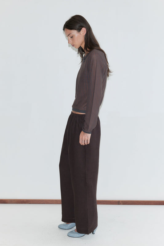 A woman with long dark hair stands in profile against a plain white background, looking down, wearing the Linen Ease Trouser - Brown Black Stripe by Deiji Studios, dark brown fine-striped wide-leg linen trousers, with a matching brown striped knit top and pale blue heels.