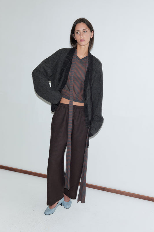 A woman with long dark hair stands against a plain white background wearing the Linen Ease Trouser - Brown Black Stripe by Deiji Studios, dark brown fine-striped wide-leg linen trousers, with a brown striped knit top, a charcoal cardigan and pale blue heels.