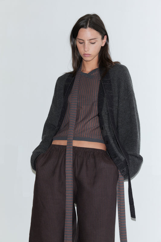 A cropped view of a woman wearing the Linen Ease Trouser - Brown Black Stripe by Deiji Studios, dark brown fine-striped wide-leg linen trousers, with a brown striped knit top and an open charcoal cardigan against a plain white background.