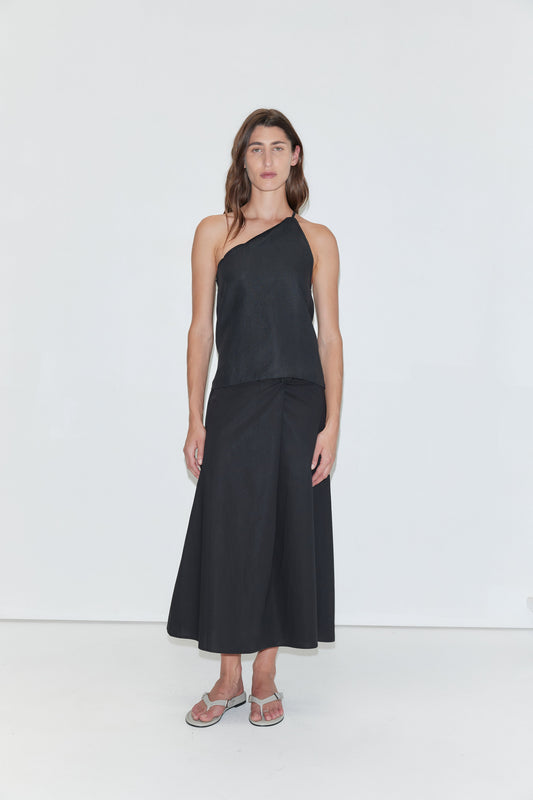 A person stands against a plain white background, wearing the Deiji Studios Half Halter - Black linen one-shoulder top, a long black skirt, and silver sandals. They have long brown hair and a neutral expression.