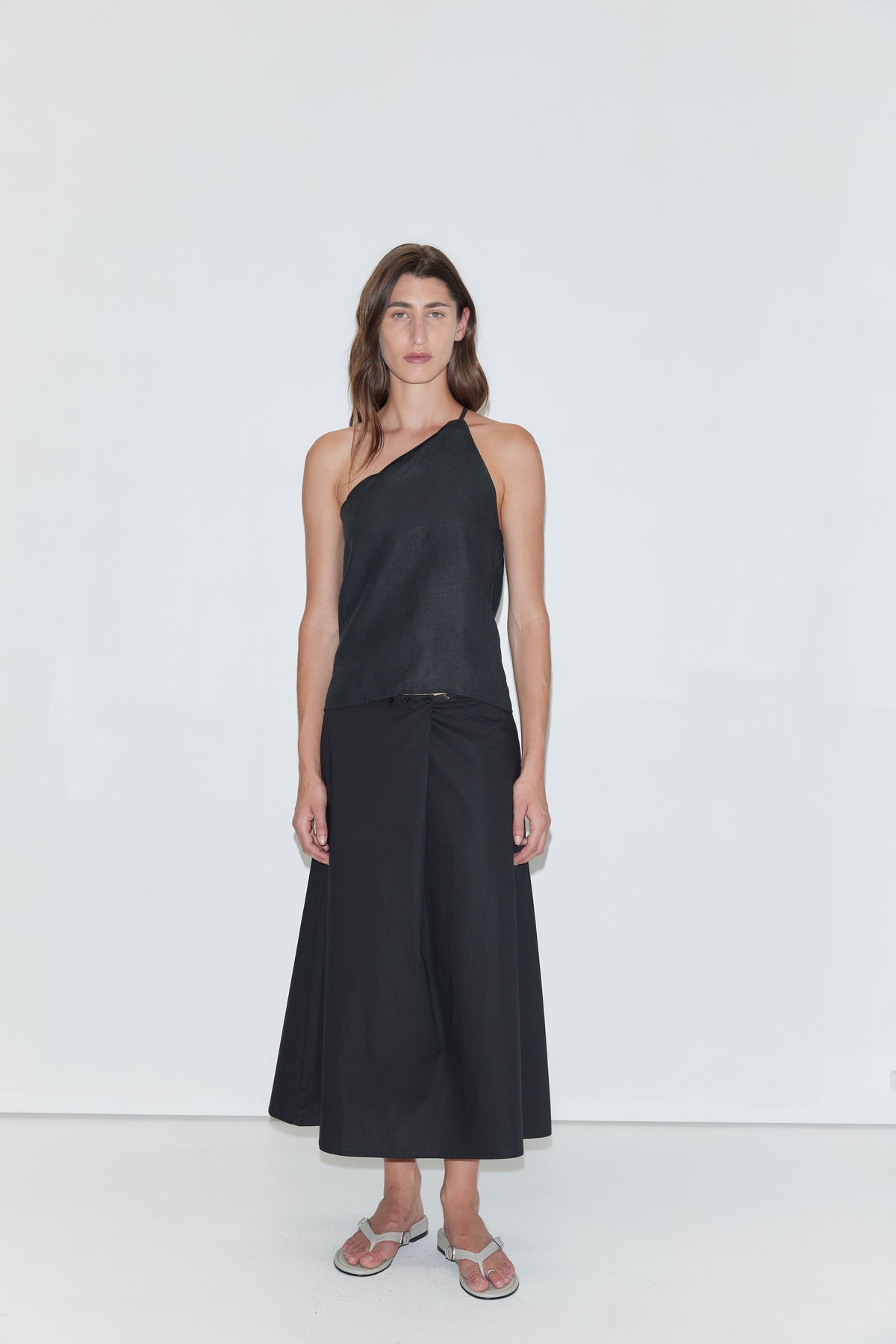 A person stands against a plain white background, wearing a black one-shoulder top and the Wrap Skirt - Black from Deiji Studios, paired with sandals.