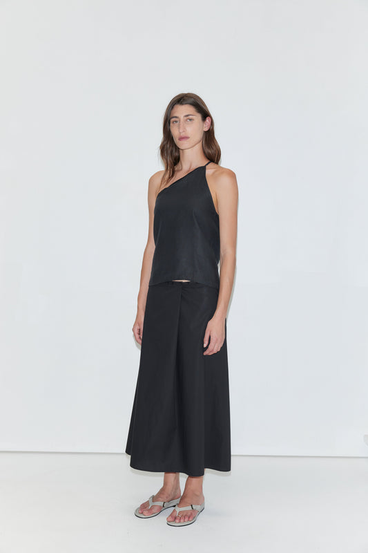 A woman stands against a white background wearing a black one-shoulder top, the Wrap Skirt - Black from Deiji Studios in cotton poplin with an A-line silhouette, and silver flip-flops. She has long brown hair and a neutral expression.