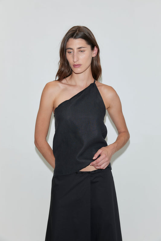 A woman with long brown hair wears the Wrap Skirt - Black by Deiji Studios, a sleeveless one-shoulder black top, and stands against a plain white background.
