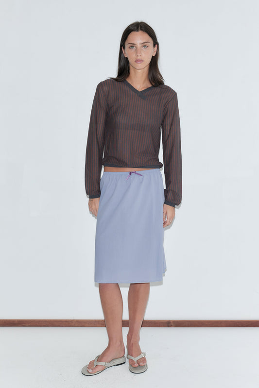 A woman with long dark hair stands against a plain white background wearing the Stripe Longsleeve - Brown Grey Stripe by Deiji Studios, a sheer brown and charcoal striped knit V-neck top, with a periwinkle midi skirt and flip-flops.