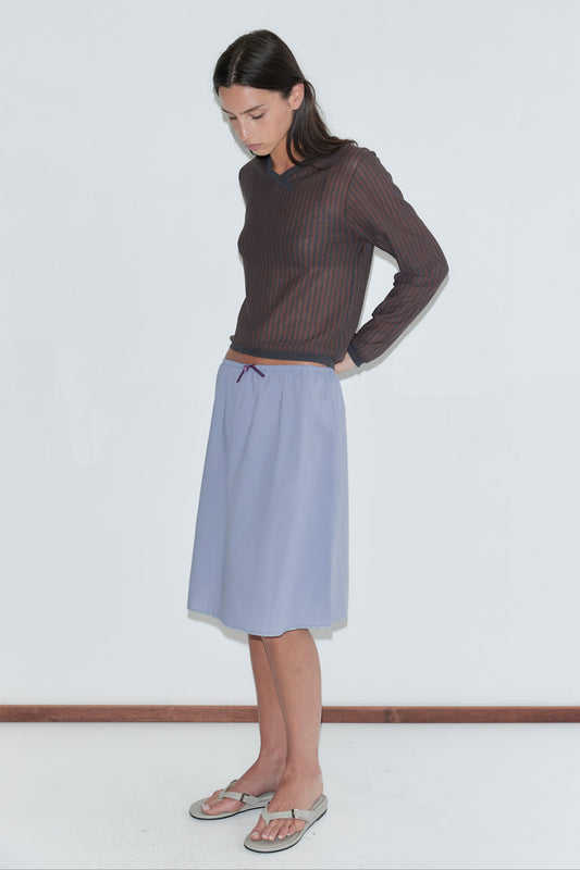 A woman with long dark hair stands in profile against a plain white background wearing the Stripe Longsleeve - Brown Grey Stripe by Deiji Studios, a sheer brown and charcoal striped knit top, with a periwinkle midi skirt and flip-flops.