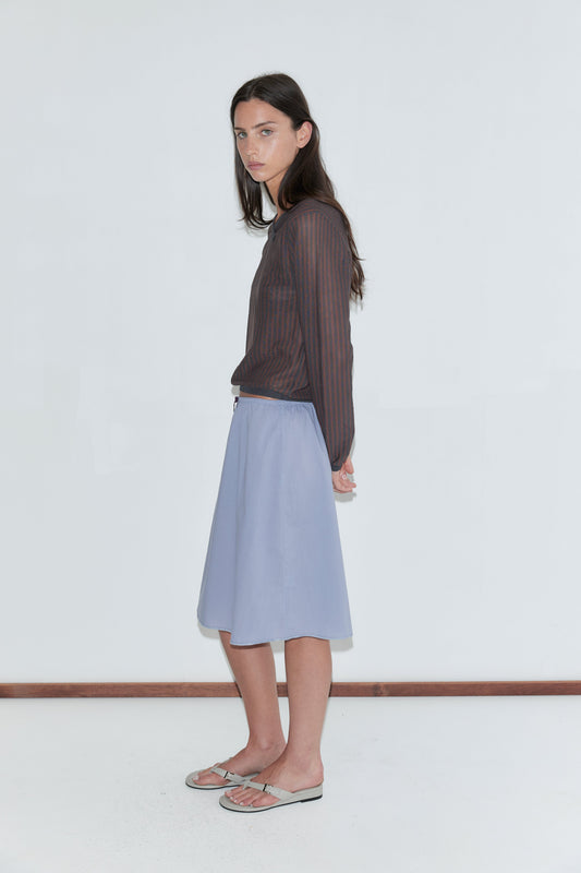 A woman with long dark hair stands in profile against a plain white background wearing a sheer brown striped long-sleeve top and the Basic Skirt - Thistle by Deiji Studios, a periwinkle cotton voile midi skirt with an elasticated waist, and grey flip-flops.
