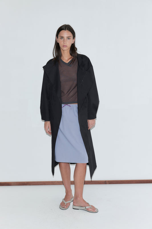 A woman with long dark hair stands against a plain white background wearing a long open black coat over a sheer brown striped top and the Basic Skirt - Thistle by Deiji Studios, a periwinkle cotton voile midi skirt, with grey flip-flops.