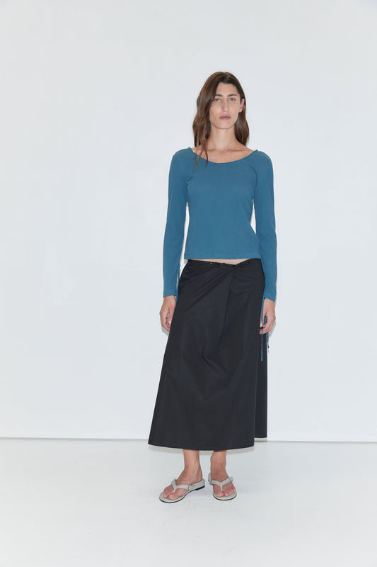 A person stands against a plain white background, wearing the Cuff Tie Top - Teal by Deiji Studios, paired with a long black skirt and sandals.