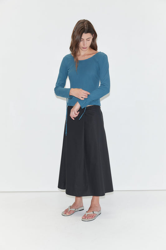 A person stands against a white background, wearing the Cuff Tie Top - Teal from Deiji Studios styled with a black wide-leg skirt and gray flip-flops, looking down with one hand touching their waist.