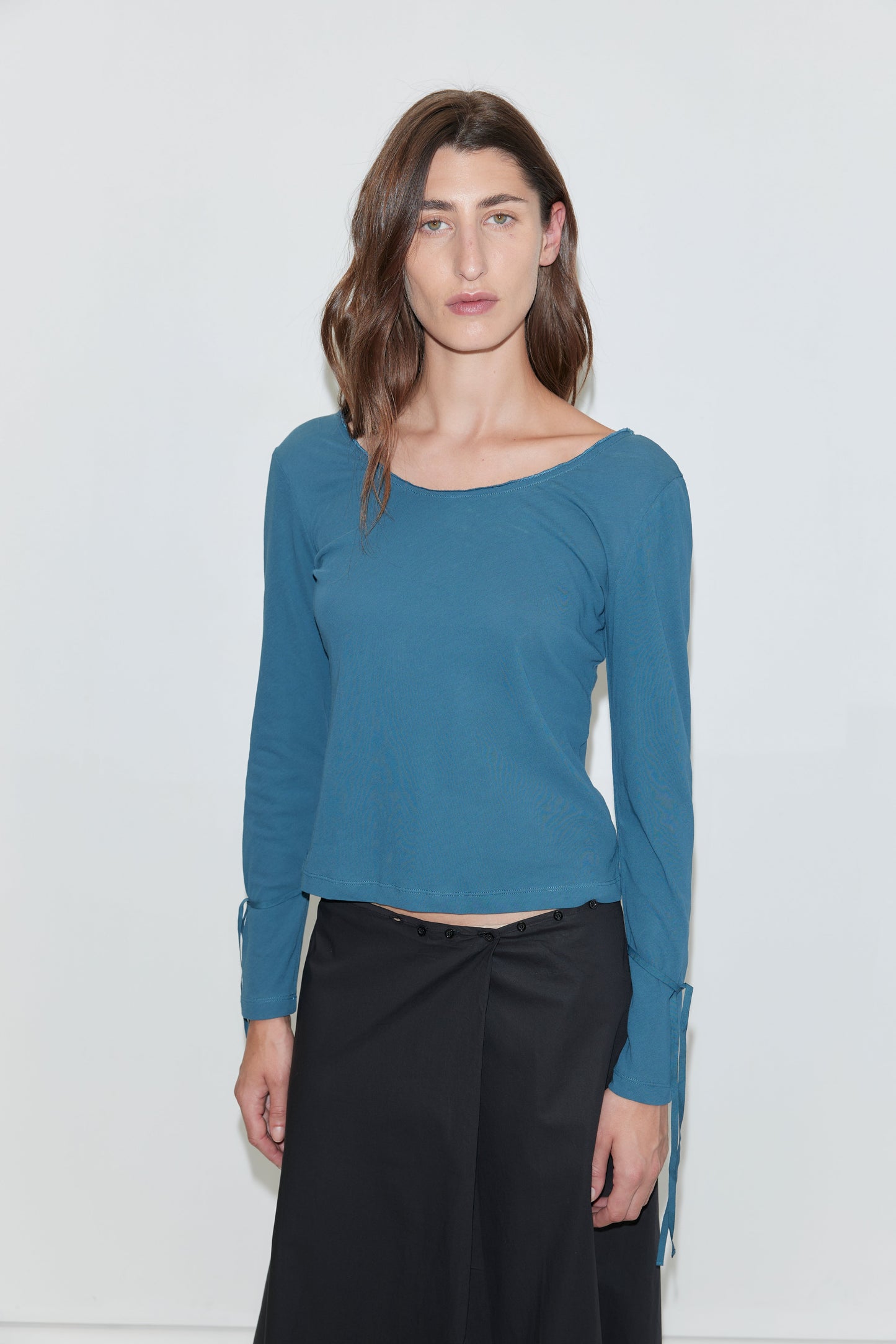 A woman with wavy brown hair stands against a white background, wearing the Cuff Tie Top - Teal from Deiji Studios, featuring long sleeves with small side ties at the cuffs, paired with a black button-front skirt. She faces forward with a neutral expression.