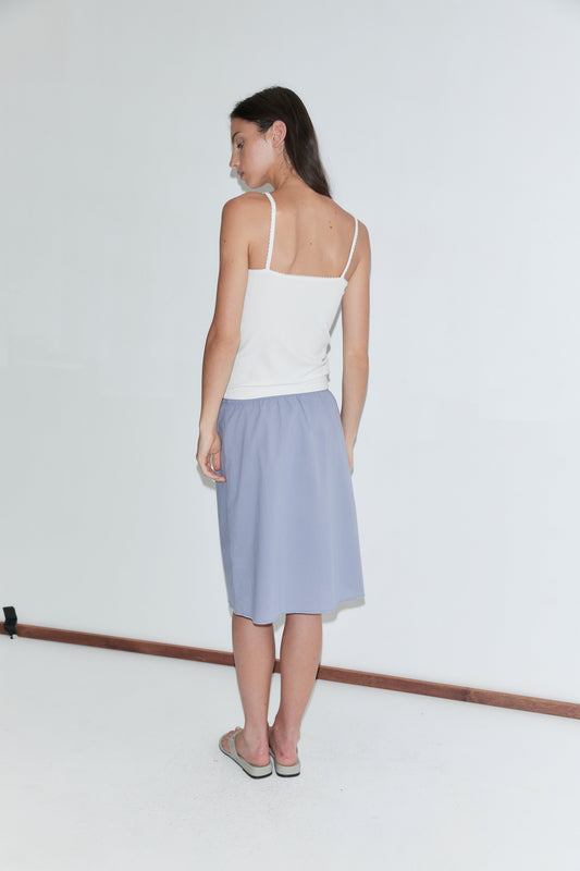 A woman with long dark hair stands with her back to the camera against a plain white background, wearing a white scallop-strap tank and the Basic Skirt - Thistle by Deiji Studios, a periwinkle cotton voile midi skirt, with grey flip-flops.