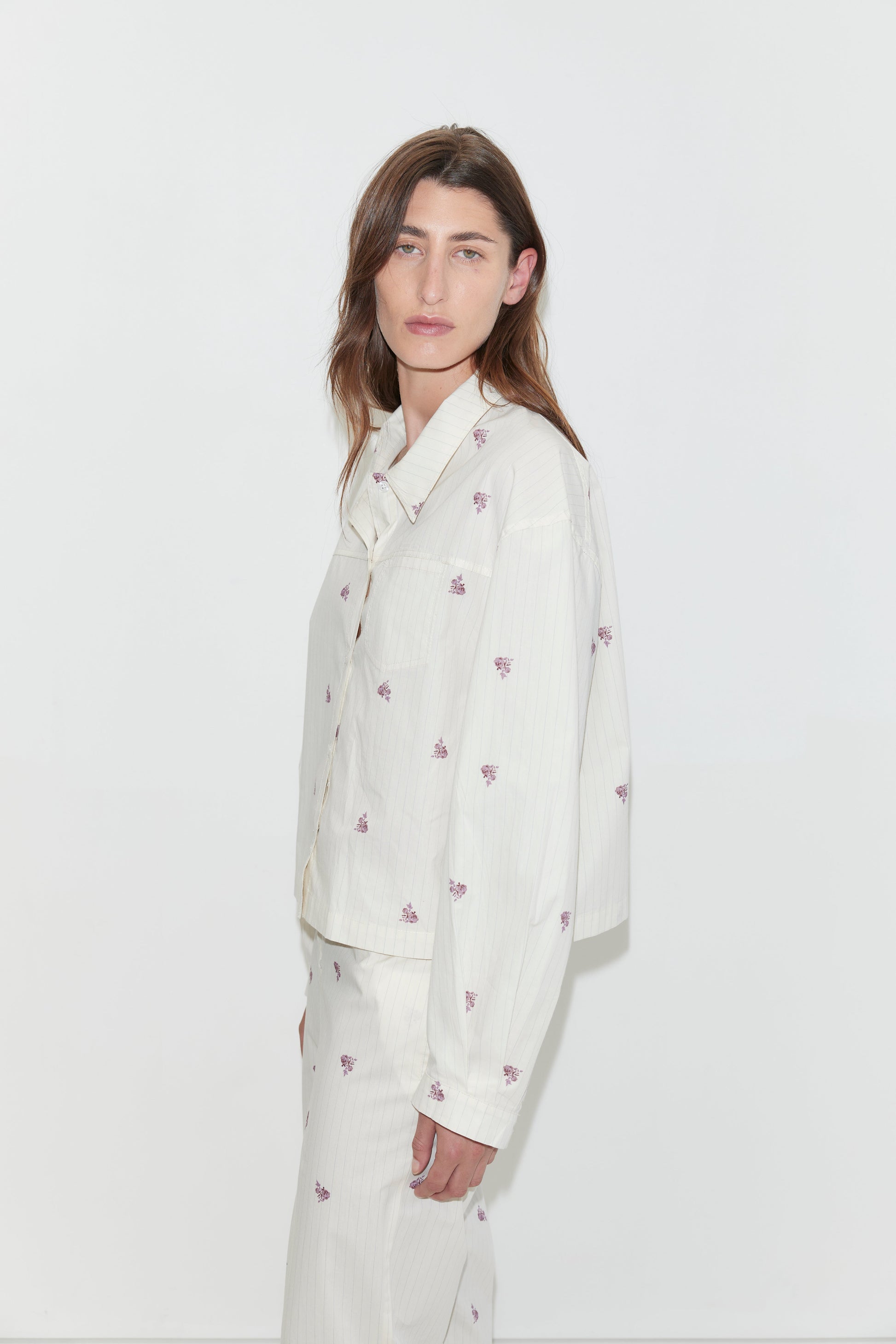 A person with long brown hair, wearing an oversized light-colored organic cotton Boxy Shirt - Floral Stripe and matching pants with small purple floral prints, stands against a plain white background, looking slightly toward the camera.