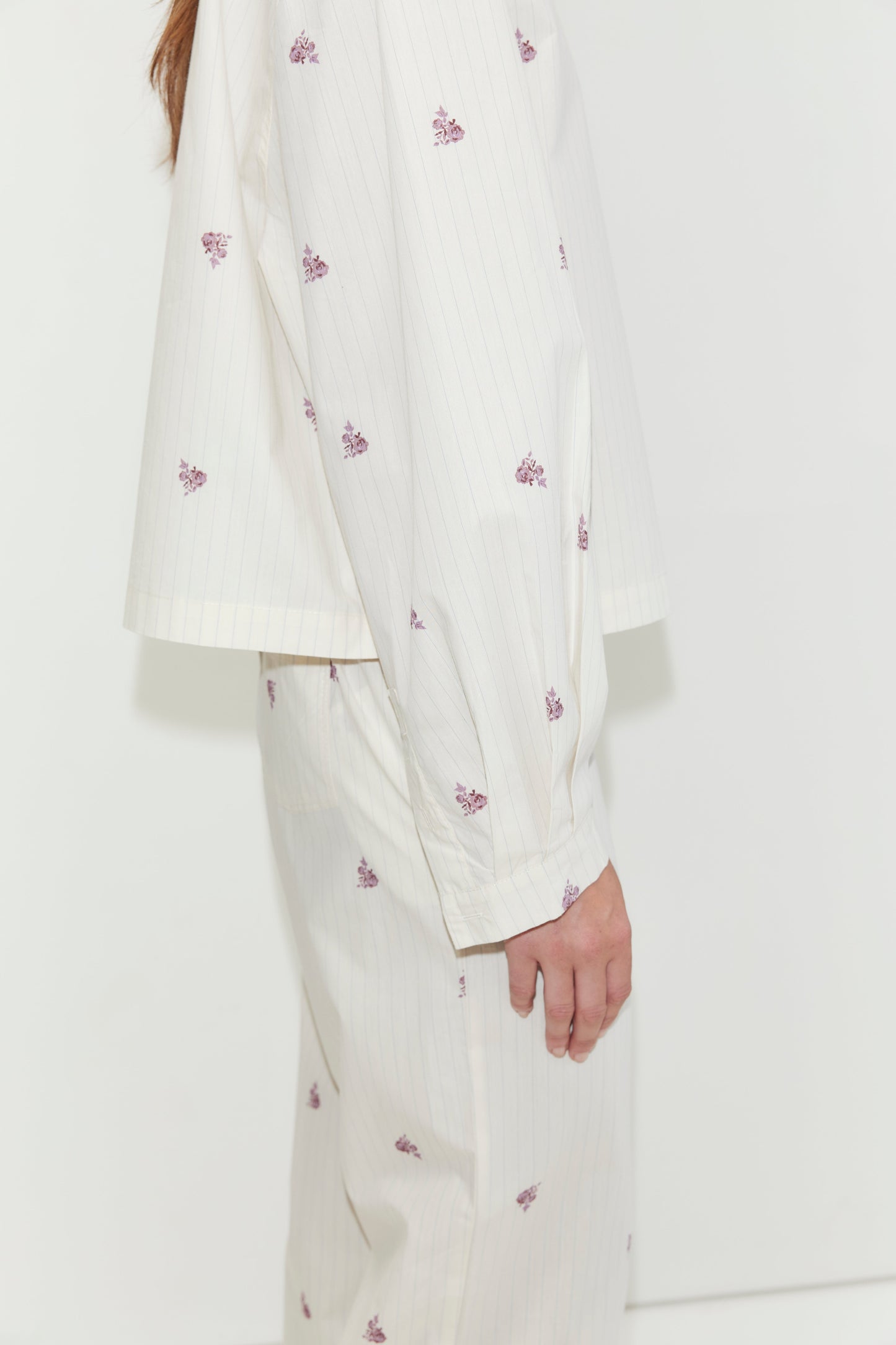 A person stands sideways against a plain white background, wearing the Boxy Long Set - Floral Stripe. This cream-colored pajama set features thin stripes and small purple floral prints, and is crafted from cotton poplin for a relaxed, oversized fit with a matching shirt and bottoms.