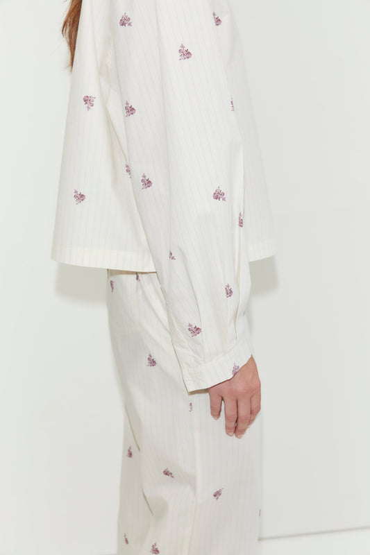 A person stands sideways against a plain white background, wearing the Boxy Long Set - Floral Stripe. This cream-colored pajama set features thin stripes and small purple floral prints, and is crafted from cotton poplin for a relaxed, oversized fit with a matching shirt and bottoms.
