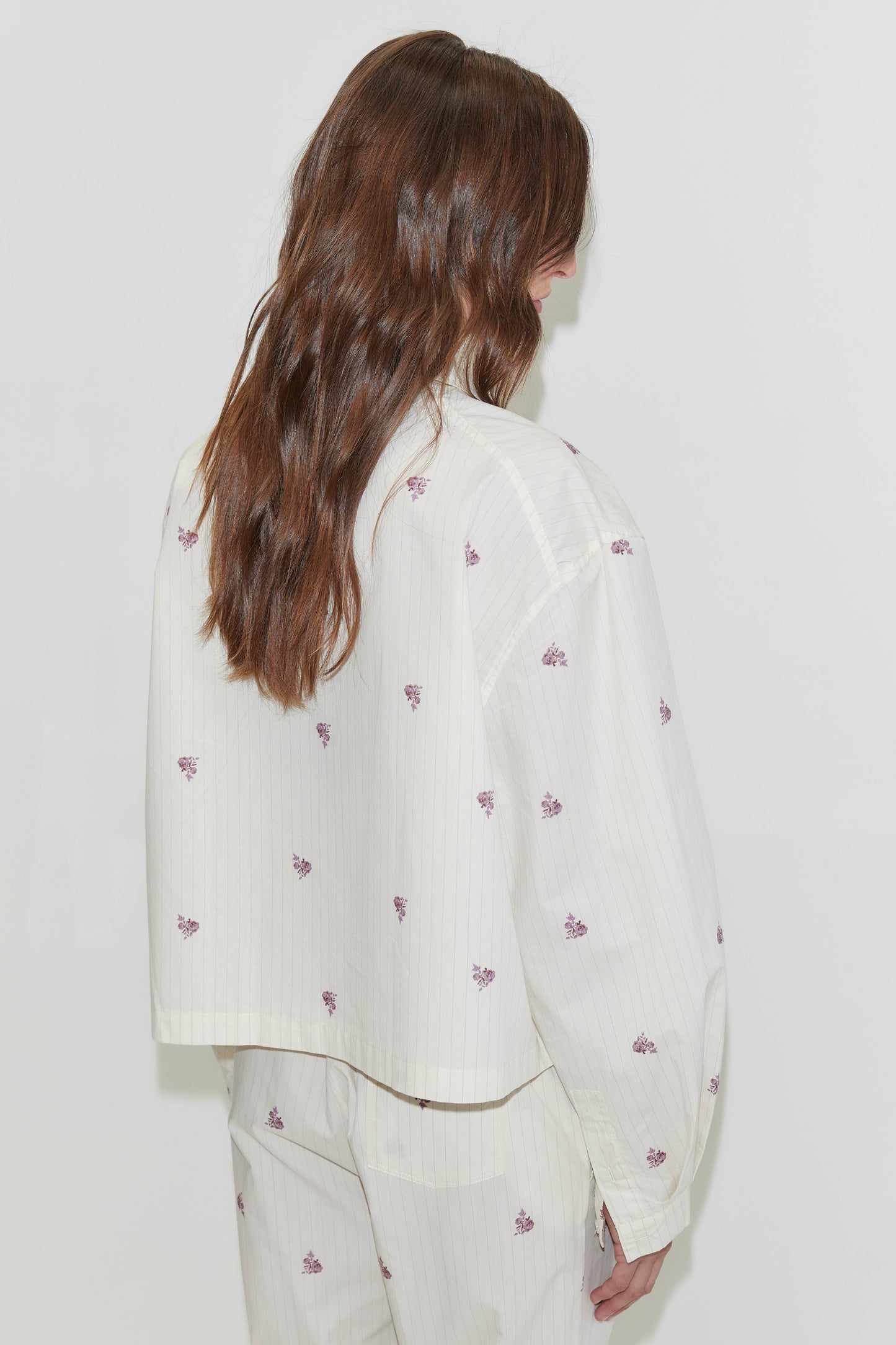 A person with long brown hair stands facing away, wearing the Boxy Shirt - Floral Stripe, an organic cotton shirt adorned with small purple floral patterns, paired with matching pants against a plain white background.