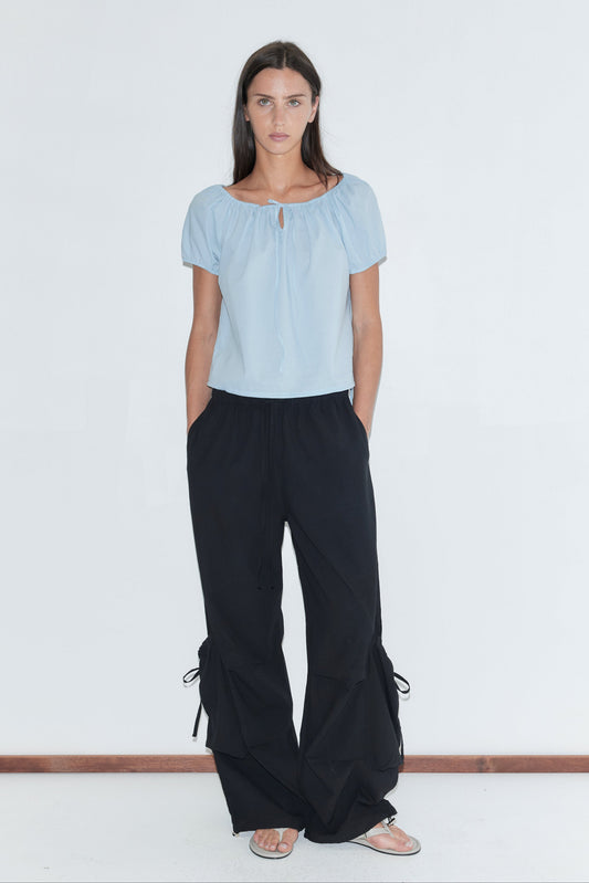 A woman with long dark hair stands against a plain white background wearing the Tie Top - Blue by Deiji Studios, a pale blue top with a gathered neckline and puff sleeves, with black wide-leg trousers and flip-flops.