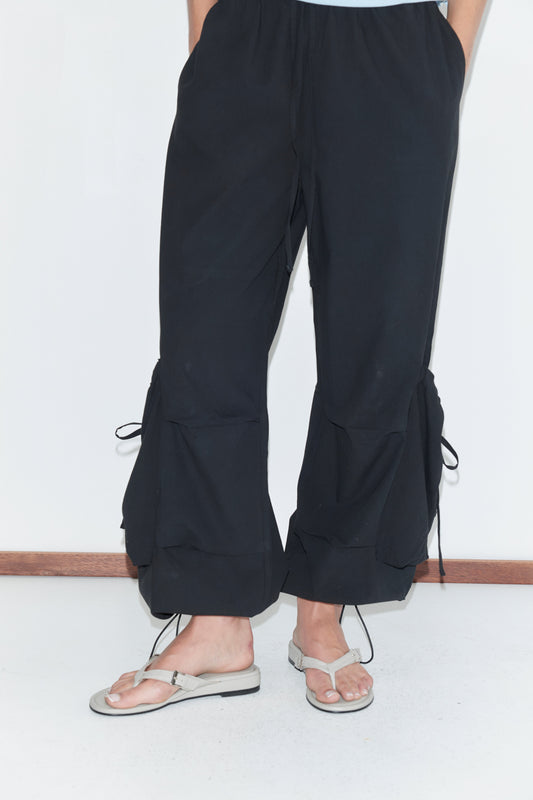 A cropped view of the Drawcord Pocket Pant - Black by Deiji Studios, black wide-leg trousers with side pockets and drawcord ties gathering the lower legs, worn with flip-flops against a plain white background.