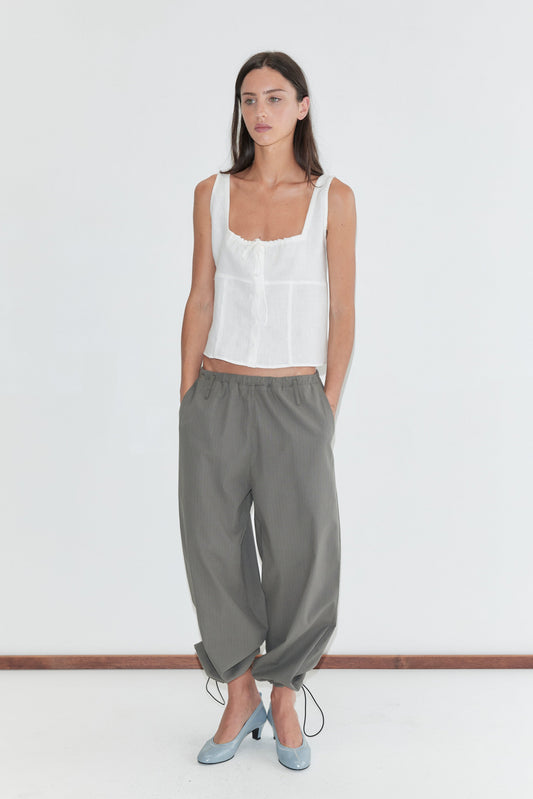 A woman with long dark hair stands against a plain white background wearing the Relax Tie Top - White by Deiji Studios, a white sleeveless linen top with a gathered drawstring neckline, with grey check trousers and pale blue heels.