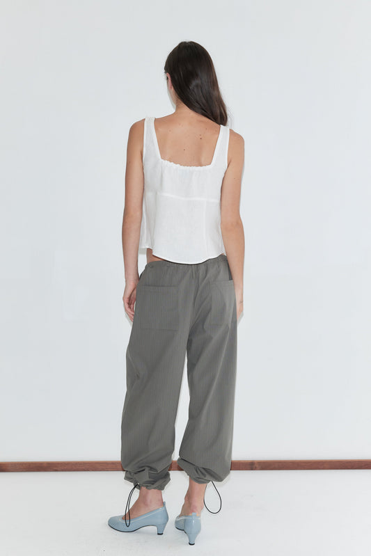 A woman with long dark hair stands with her back to the camera against a plain white background wearing the Relax Tie Top - White by Deiji Studios, a white sleeveless linen top, with grey check trousers and pale blue heels.