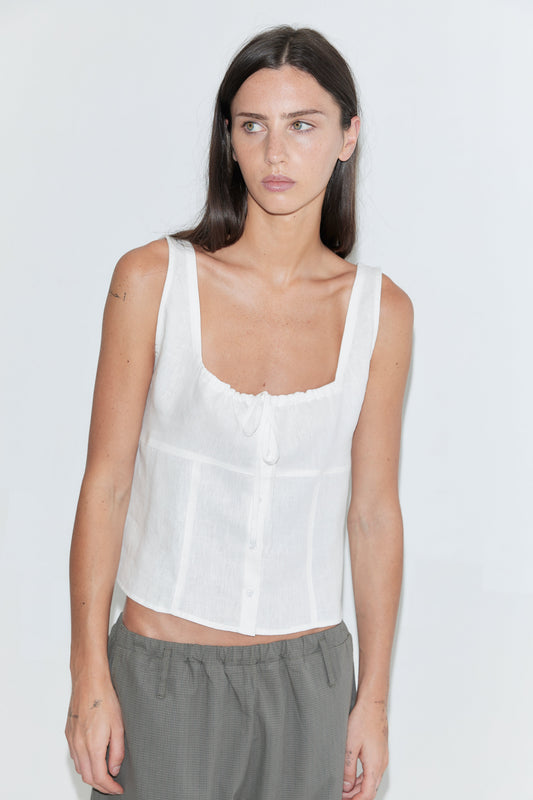 A cropped front view of the Relax Tie Top - White by Deiji Studios, a white sleeveless linen top with wide straps and a gathered drawstring tie at the neckline, worn with grey trousers against a plain white background.