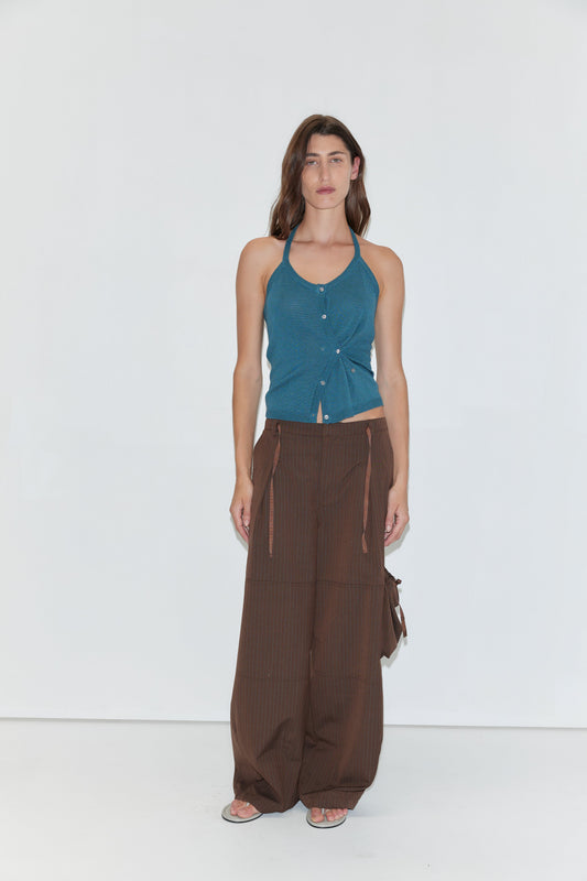 A person stands against a white background wearing the Cross Top - Aqua, a blue fitted knit halter top with adjustable self-tie straps, paired with brown wide-leg drawstring pants and sandals.