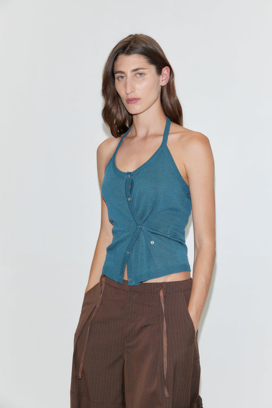 A woman with long brown hair wears the Cross Top in Aqua—a fitted knit halter top with adjustable self-tie straps—paired with brown pants, standing against a plain white background and looking at the camera with a neutral expression.