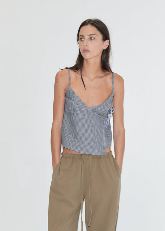 A woman with long dark hair stands against a plain white background wearing the Wrapping Top - Indigo Check by Deiji Studios, an indigo micro-check linen cami with a wrap front and side tie, with khaki drawcord trousers.