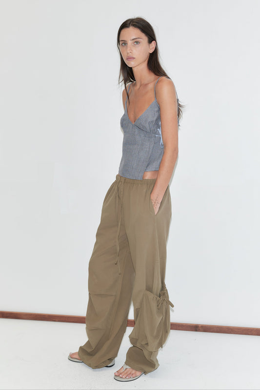 A woman with long dark hair stands in profile against a plain white background wearing the Drawcord Pocket Pant - Khaki by Deiji Studios, khaki wide-leg trousers, with a blue check cami top and flip-flops.