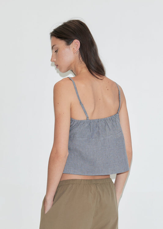 A woman with long dark hair stands with her back to the camera against a plain white background wearing the Wrapping Top - Indigo Check by Deiji Studios, an indigo micro-check linen cami with a gathered back, with khaki trousers.