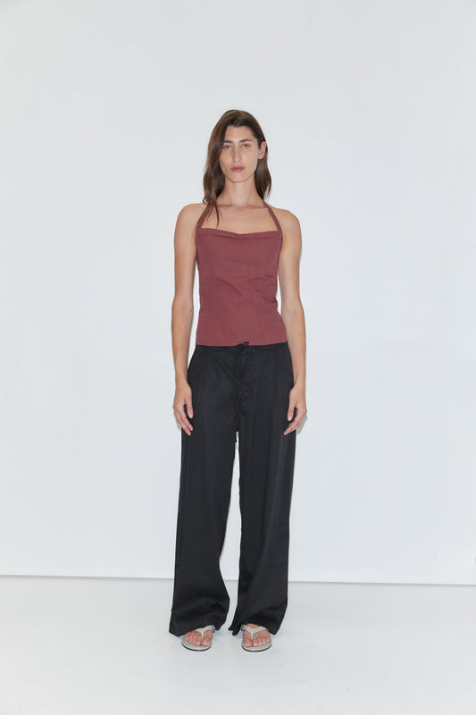 A person stands against a plain white background wearing a sleeveless maroon top, loose wide-leg Tencel Pant in Black by Deiji Studios, and sandals. They have long brown hair and a neutral expression, with arms relaxed at their sides.