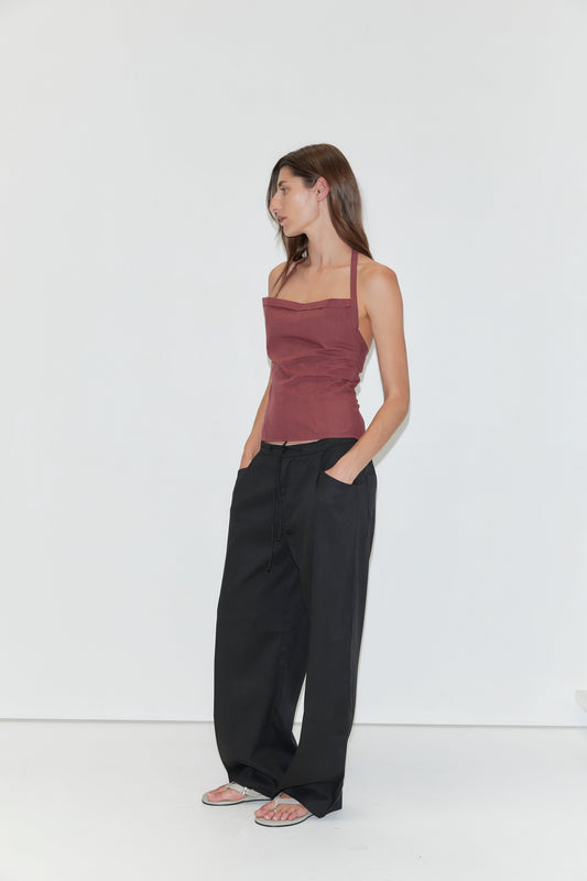 A woman stands against a white background, wearing the Folded Top in Rust Stripe, paired with loose black pants and sandals. She has long brown hair and looks to her left with her hands in her pockets.