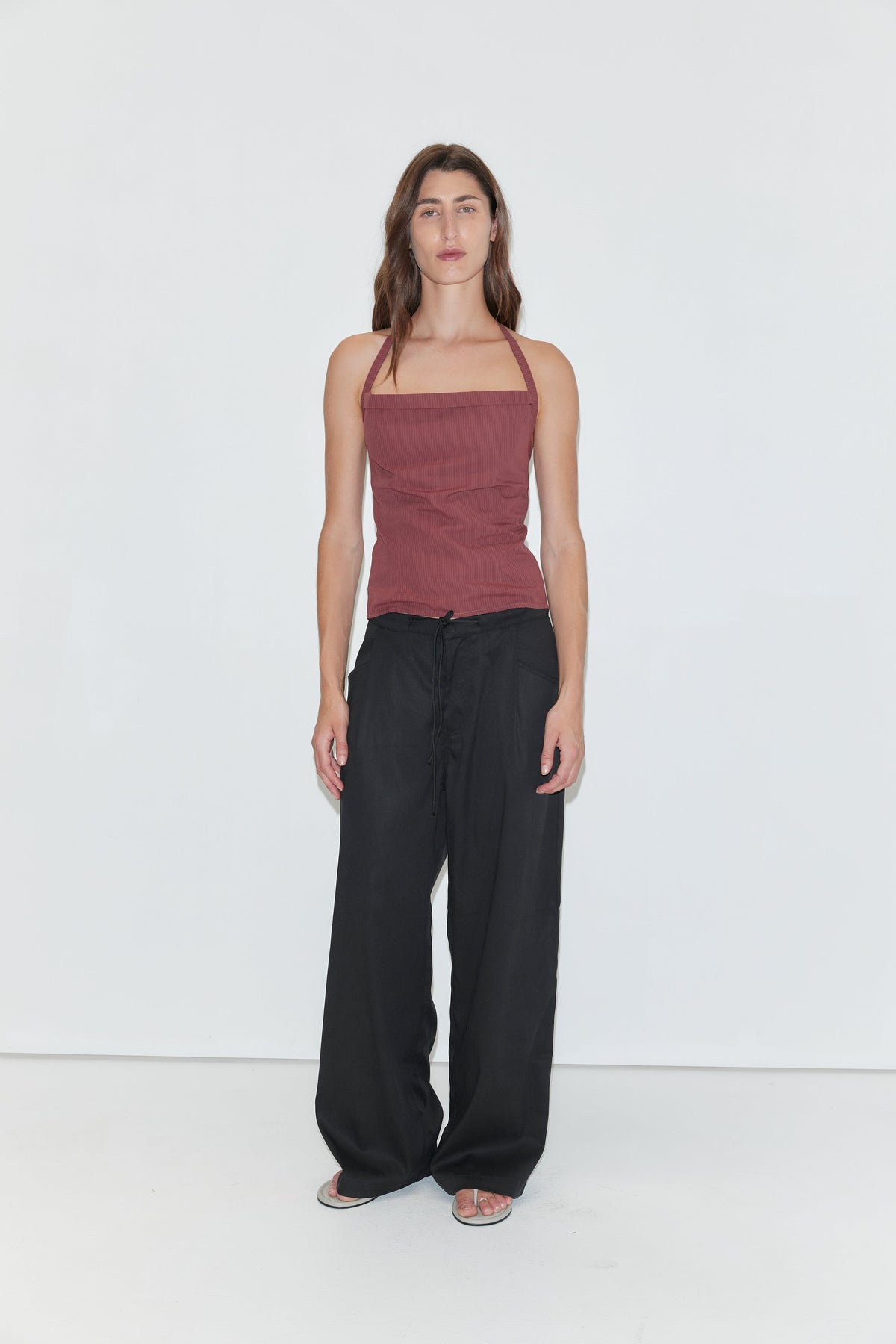 A person with long brown hair stands against a plain white background, wearing the Folded Top in Rust Stripe by Deiji Studios, paired with loose black pants and sandals.