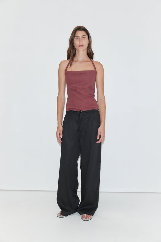 A person with long brown hair stands against a plain white background, wearing the Folded Top in Rust Stripe by Deiji Studios, paired with loose black pants and sandals.
