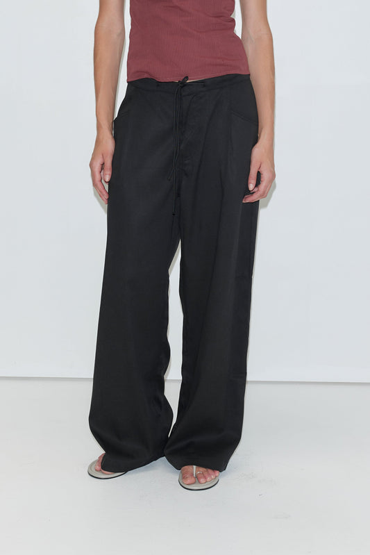A person wearing a sleeveless maroon top, loose black Tencel Pant - Black from Deiji Studios, and white flip-flops stands against a plain white background.